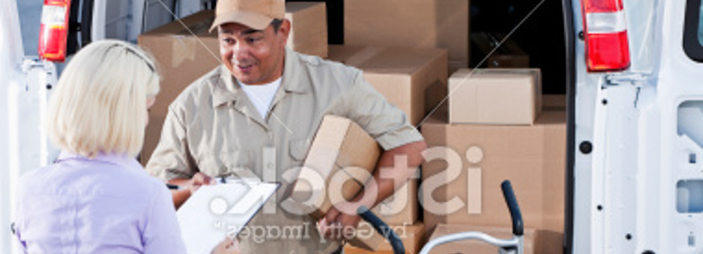Delivery person holding out clipboard for lady to sign, both standing at the back of his box filled truck